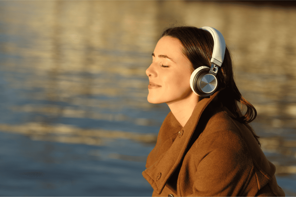 Lady wearing noise cancelling headphones smiling with her eyes closed by a lake.